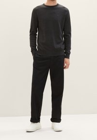 Suéter gris de punto con un cuello redondo, combinado con pantalones negros y zapatillas blancas, posando frente a un fondo neutro.