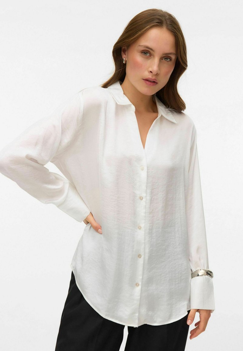 White, long-sleeve button-up shirt with a relaxed fit, soft texture, and a collar. Button detailing and a curved hem. Worn with black pants.