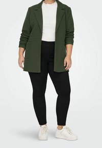 Zelený strukturovaný blazer s klopami a dvěma předními kapsami, spárovaný s černými úzkými kalhotami a bílými teniskami. Pod ním jednoduchý bíle proužkovaný top.