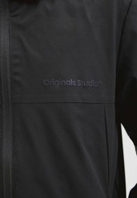 Veste noire avec fermeture éclair verticale et logo "Originals Studio™" imprimé sur le côté gauche de la poitrine.