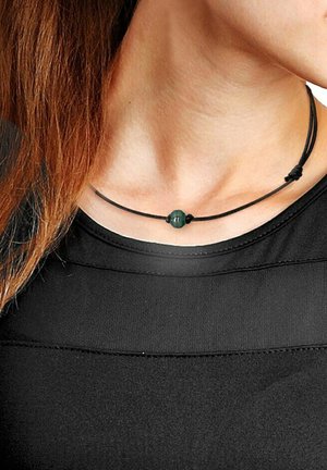 Collier en cordon noir avec une perle centrale verte, de forme circulaire, texture lisse, et sécurisé par deux nœuds. Porté près du cou.