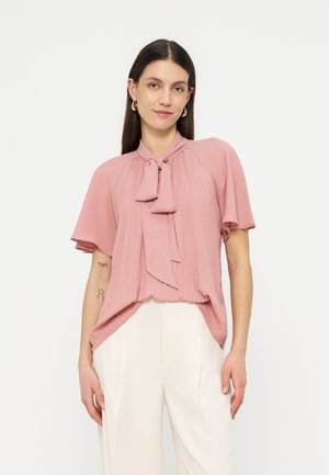 Woman wearing a pink pleated blouse with a bow tie collar and white high-waisted pants, standing against a plain white background.