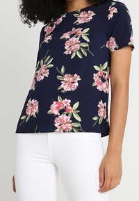Short-sleeve navy blouse with a floral print in pink and green, featuring a loose fit and a smooth texture. Paired with white pants.