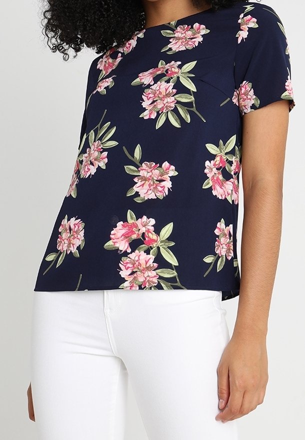 Short-sleeve navy blouse with a floral print in pink and green, featuring a loose fit and a smooth texture. Paired with white pants.