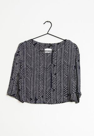 Blouse noire à manches longues avec motif de lignes diagonales blanches, col rond, suspendue sur un cintre noir sur fond blanc.