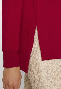 Suéter rojo con puños acanalados y una abertura lateral, con bolsillos estructurados. La tela parece suave y tiene una textura lisa.