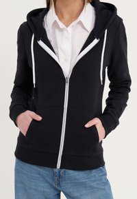 Femme portant un sweat à capuche noir zippé avec des cordons blancs sur une chemise blanche à col et un jean bleu, mains dans les poches du sweat.