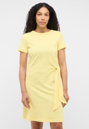 Frau mit lockigem Haar, die ein knielanges, gelbes Kleid mit kurzen Ärmeln und einem seitlichen Knotendetail vor einfarbigem Hintergrund trägt.