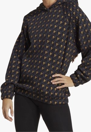 Frau trägt schwarzen Hoodie mit goldenen Adidas-Logos und schwarzen Leggings, Hand in der Hüfte, zugeschnitten bei Hals und Oberschenkeln.