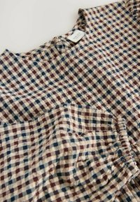 Blusa a quadri nei colori beige, navy e bordeaux. Tessuto morbido con scollatura tonda, presenta arricciature lungo i bordi e un'etichetta sul retro.