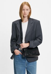 Borsa a spalla in pelle nera con texture liscia, chiusura a patta con dettagli a pulsante e tracolla regolabile, abbinata a un blazer grigio a righe.