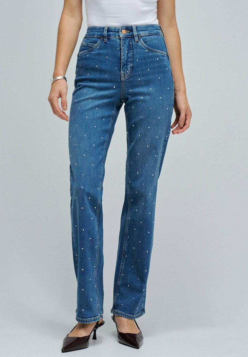 Mujer con pantalones vaqueros de cintura alta azules con pequeños lunares blancos, combinados con tacones negros puntiagudos y una blusa sin mangas blanca.