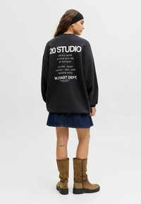 Černá oversized mikina s bílým textem na zádech, spárovaná s džínovou sukní a hnědými kozačkami po kolena. Hladká látka, uvolněný střih.