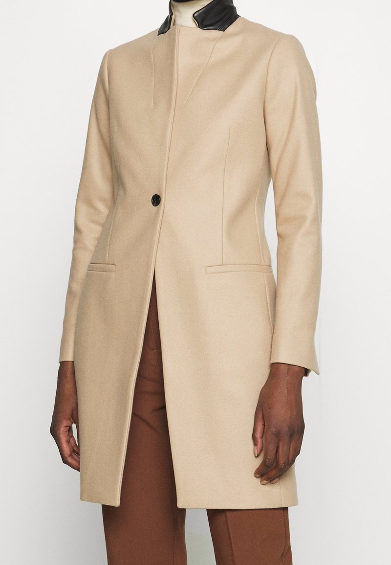 Manteau long beige avec un bouton noir, col noir, coupe droite, porté par-dessus un pantalon marron sur une personne à la peau foncée.