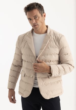 Homme portant une veste matelassée beige par-dessus une chemise blanche, boutonnant la veste, debout devant un fond clair uni.