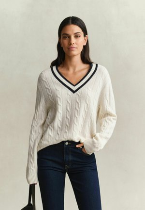 Femme aux longs cheveux foncés portant un pull col en V blanc à torsades avec des bordures foncées et un jean bleu foncé, tenant un sac noir.