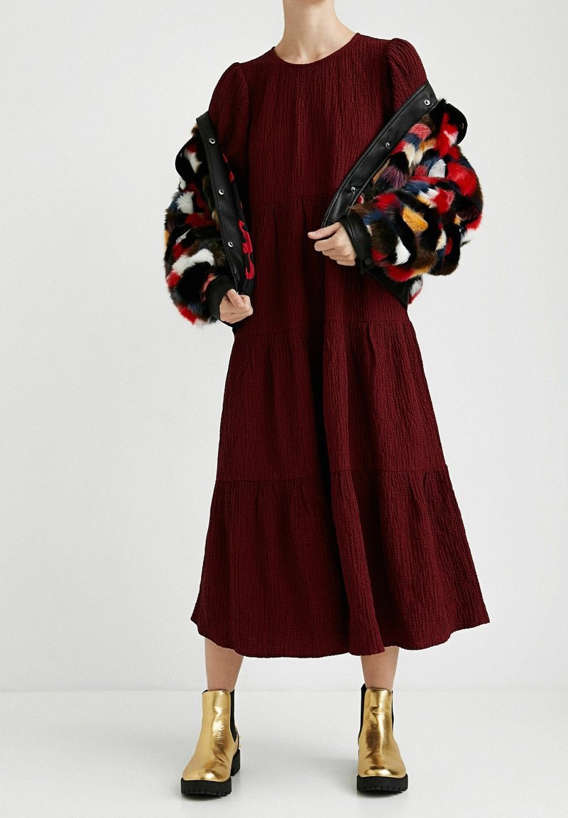 Robe bordeaux à volants en tissu texturé, associée à une veste en fausse fourrure colorée et à des bottines dorées métalliques avec une semelle chunky.