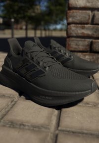 Pár černých běžeckých bot Adidas s podrážkou Boost na zpevněném venkovním povrchu poblíž kamenné zdi za denního světla.