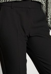 Czarne spodnie o dopasowanym kroju z gładkiej tkaniny, z średnim stanem i subtelnymi plisami z przodu. Noszone z wzorzystą, lekką bluzką.