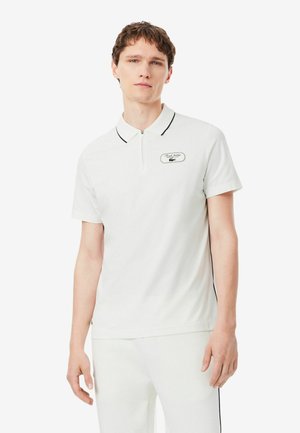 Giovane uomo con capelli corti e ricci indossa una polo bianca con un piccolo logo ricamato e rifinitura nera sul colletto.