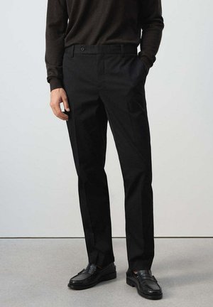 Hombre con pantalones negros a medida, mocasines de cuero negro y un suéter marrón oscuro de manga larga, de pie con una mano en el bolsillo.