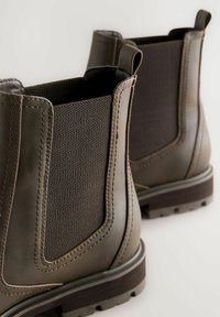 Ett par bruna läder Chelsea-boots med elastiska sidopaneler och dragflikar, visade i närbild mot en enkel bakgrund.