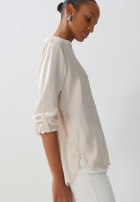 Blusa beige chiaro con colletto alla mandarino, maniche lunghe arrotolate fino al gomito, spacchi laterali e tessuto morbido e fluido. Abbinata a pantaloni bianchi.
