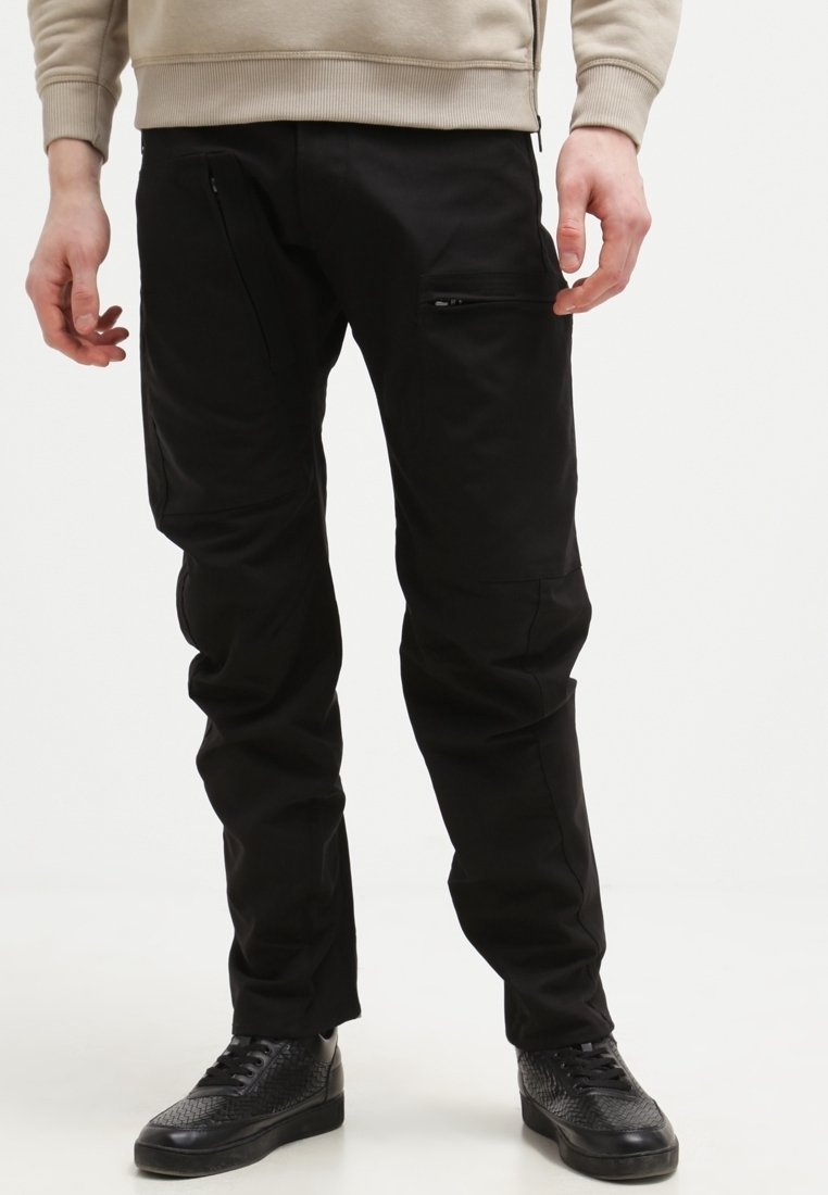 Svarta byxor med en slät tygkvalitet och flera fickor med dragkedja. Rakt bensnitt, med subtila sömmar och en modern passform. Texturerade svarta skor.