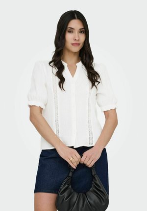 Femme aux longs cheveux noirs portant un chemisier blanc à manches bouffantes et une jupe bleu marine, tenant devant elle un sac à main en cuir noir froncé.