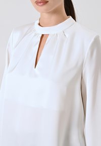 Weiße Bluse mit hohem Ausschnitt und Schlitz, gefälteter Gestaltung und glatter, satinähnlicher Textur. Die langen Ärmel sind eng geschnitten.