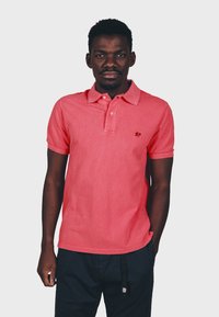 Korallenfarbenes Poloshirt mit klassischem Kragen, versehen mit einem kleinen Elefantenlogo auf der Brust; kombiniert mit dunklen Shorts und einem strukturierten Stoff.