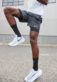 Șorturi atletice gri cu un căptușel negru, asortate cu adidași Nike albi și gri și șosete negre, prezentând o poziție dinamică pe o suprafață texturată.
