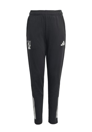 Jong persoon in blauwgroen en zwart voetbaltenue met een zwart-witte voetbal, glimlachend, met witte voetbalschoenen tegen een effen achtergrond.