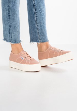 Osoba nosząca jasnoróżowe sneakersy na platformie z białymi podeszwami i jasnoniebieskie dżinsy z postrzępionym dołem na białej podłodze.