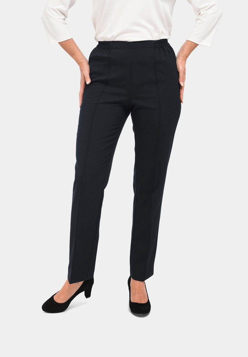 Zwarte broek met een tailored ontwerp, elastische tailleband, rechte pijpen en subtiele voornaden. Gecombineerd met zwarte hakken tegen een lichte achtergrond.