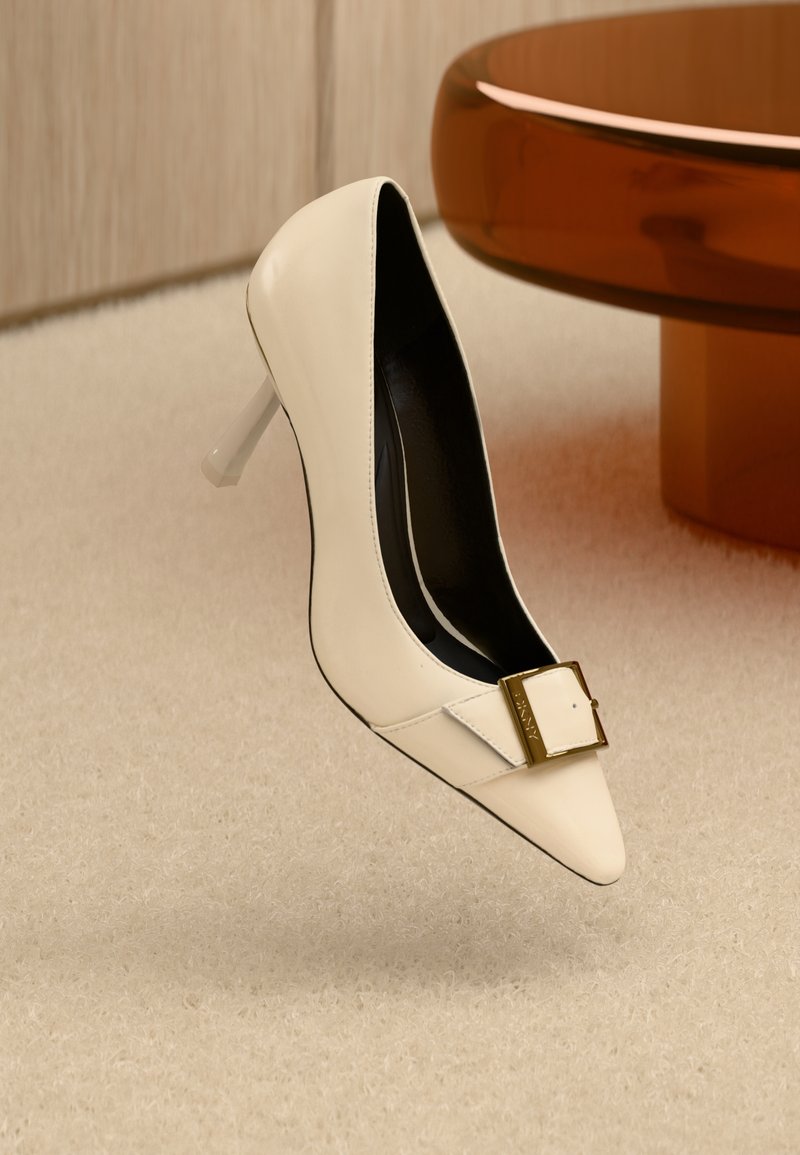 White leather high-heeled shoe with a pointed toe, clear plastic heel, and a gold buckle accent. Black interior lining.