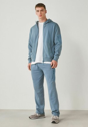 Young man standing wearing light blue zip-up hoodie, matching cargo pants, white T-shirt, and gray sneakers against plain background.
