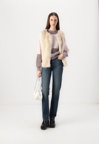 Un gilet en fausse fourrure claire sur un pull rayé dans des tons de rose et de violet. Jeans bleu foncé et chaussures à plateforme noires, tenant un sac à main blanc.