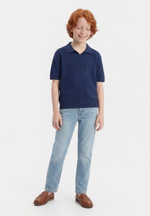 Červenovlasý chlapec sa usmieva, má na sebe tmavomodré polo tričko s krátkym rukávom, svetlomodré rifle a hnedé mokasíny, stojí pred jednoduchým bielym pozadím.