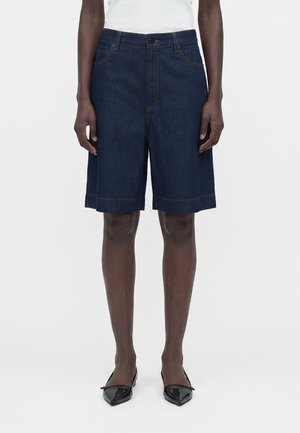 Άτομο που φορά σκούρες μπλε τζιν σορτς μέχρι το γόνατο, λευκό τοπ και μαύρες μυτερές μπαλαρίνες, στέκεται στο φόντο λευκού χρώματος.