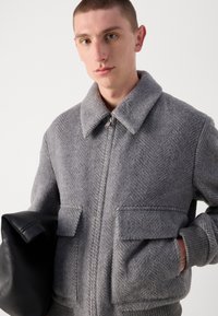 Gray herringbone jacket with a wide collar, front zip, and two large flap pockets. Model holds a black leather bag.