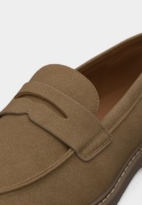 Gros plan sur un mocassin en daim marron avec un détail de bride cousue et une semelle en caoutchouc visible sur un fond blanc.