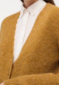 Senapsgul cardigan i fluffigt stickat material. Har en djup V-hals, ribbade ärmslut och en enda knappstängning. Bärs över en vit skjorta.