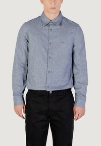 Camicia button-up azzurro chiaro in un tessuto testurizzato, con colletto classico e maniche lunghe. Indossata con pantaloni neri.