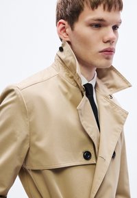 Béžový trenčkot s vysokým golierom, dvoma čiernymi gombíkmi a detailným prešívaním. Tkanina pôsobí hladko s jemným leskom.