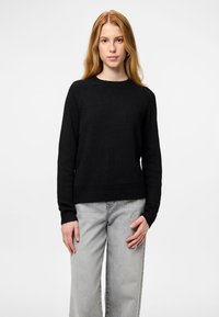 Jauna moteris su ilgiems blondiniais plaukais, vilkinti juodą megztinį ir šviesiai pilkas džinsus, stovi su neutralia veido išraiška prieš baltą foną.