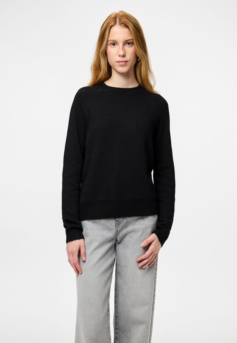 Jauna moteris su ilgiems blondiniais plaukais, vilkinti juodą megztinį ir šviesiai pilkas džinsus, stovi su neutralia veido išraiška prieš baltą foną.