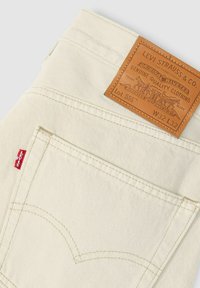 Beige Jeans aus Denim mit einem Leder-Label auf dem Bund, das das geprägte Markenlogo zeigt. Enthält einen roten Tab an der Gesäßtasche und markante Nähte.