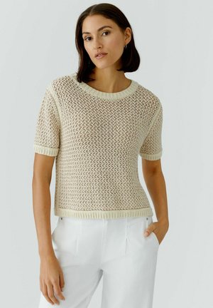 Vrouw met donker haar tot op de schouders, gekleed in een beige gebreide trui met korte mouwen en witte broek, met één hand in de zak.