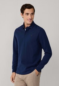 Námornícky polo s long rukávom s golierom a jemným logom na hrudi, skombinovaný s svetlohnedými štruktúrovanými nohavicami.